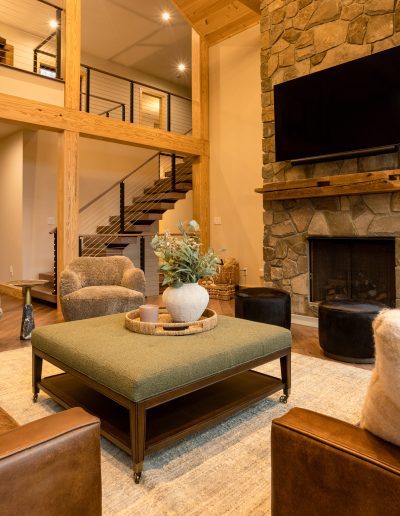 Modern living room with a stone fireplace, mounted TV, brown leather chairs, round chairs, ottomans, a green coffee table, and wooden ceiling beams. Large windows let in natural light.