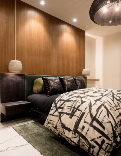 Modern bedroom with abstract decor, patterned bedding, wood accent wall, and two large colorful art pieces; grey bench and marble flooring complete the room.