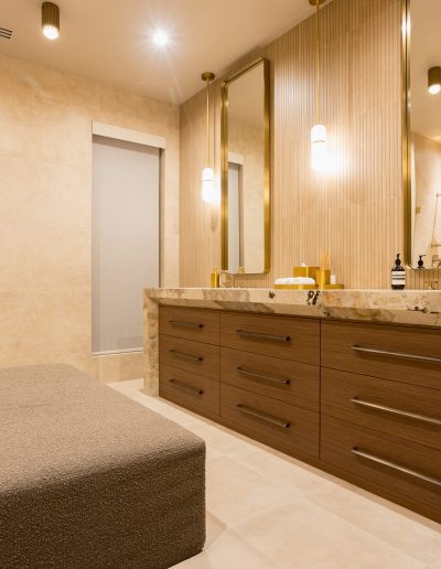 Modern bathroom with double sink vanity, marble countertops, large mirrors, wall-mounted lights, beige tiles, and a gray upholstered bench.