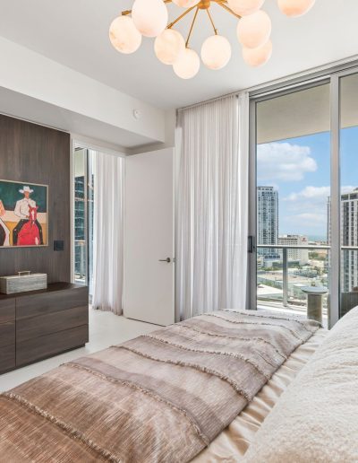 Modern bedroom with large windows, city view, neutral bedding, wood dresser, wall art, and a contemporary chandelier.